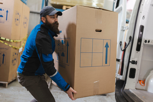 Bearded courier puts the cardboard boxes from the warehouse into the trunk of the delivery van