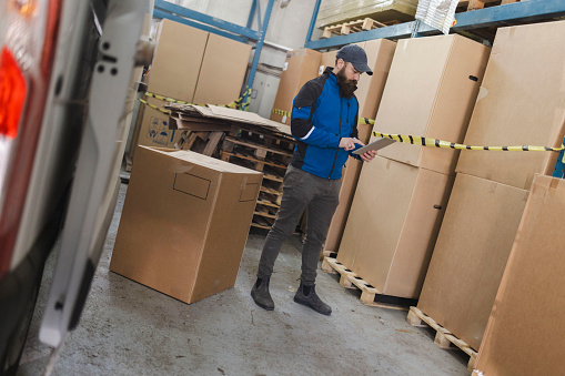 A bearded warehouse worker checks packages in a warehouse using a digital tablet