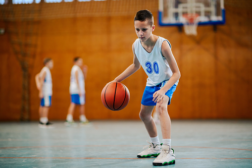 Full length of a young basketball kid dribbling and practicing basketball on court on training. In a blurry background is his junior team having training. A young athlete in action with basketball.