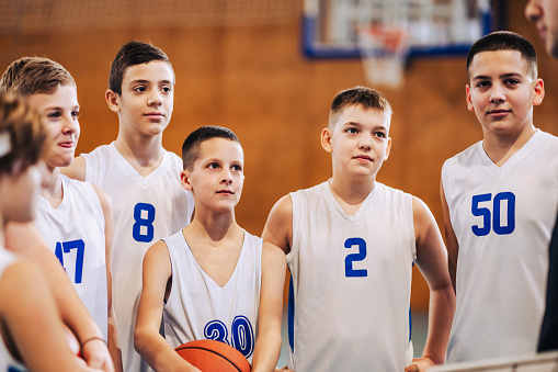 Portrait of a junior basketball team standing on court with their trainer and listening to him during training. A young basketball kids standing at indoor court and looking at the trainer on training.