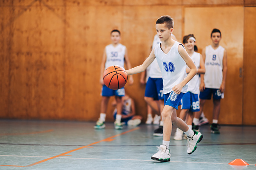 A dedicated young basketball kid is practicing and dribbling a ball on training on basketball court. In a blurry background are his teammates waiting for their turn. Junior athlete in action on court