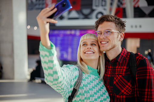 Portrait of a cheerful hipster couple standing at metro station and taking selfies while smiling at the phone. A happy urban commuters standing at train station and making self portraits with a phone