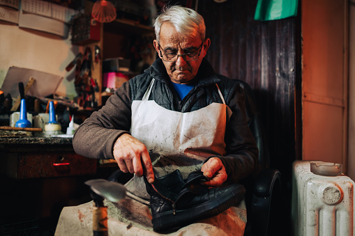 Portrait of a senior craftsman sitting at his small shoemaker workshop and working with boots. A handyman is sitting at his cobbler shop and holding boots for reparation in his hands. Craftsmanship.
