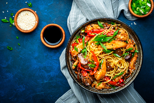 Hot stir fry egg noodles with turkey, paprika, mushrooms, chives and sesame seeds with ginger, garlic and soy sauce. Asian cuisine dish. Blue table background, top view