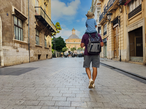 Father with kid on shoulders walk toward Basilica la Mare de Deu dels Desemparats during their family trip to Valencia, Spain