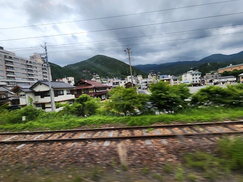 This picture appears to be taken from a moving train, given the motion blur particularly visible on the train tracks in the foreground. The scenery is of a small town or suburban area with a mix of modern and traditional buildings. The houses are relatively low-rise, with one or two stories, featuring different architectural styles and roofing materials. The backdrop is a lush, green mountainous landscape, with hills rising in the distance under a dynamic sky filled with a mix of gray storm clouds and patches of blue sky. The overall impression is of a peaceful, quiet town nestled in a verdant valley, with an impending sense of a rainstorm due to the cloudy sky.