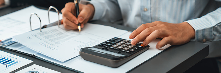 Corporate auditor calculating budget with calculator on his office desk. Dedicated accountant professional of accounting business company analyzing financial document to forecast income. Insight