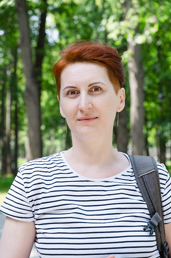Vertical portrait of a middle-aged woman with short red hair in a striped t-shirt. The woman smiles and looks into the camera. Walk in the city park.