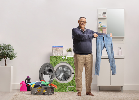 Happy mature man holding a pair of jeans in a bathroom with a power efficient washing machine
