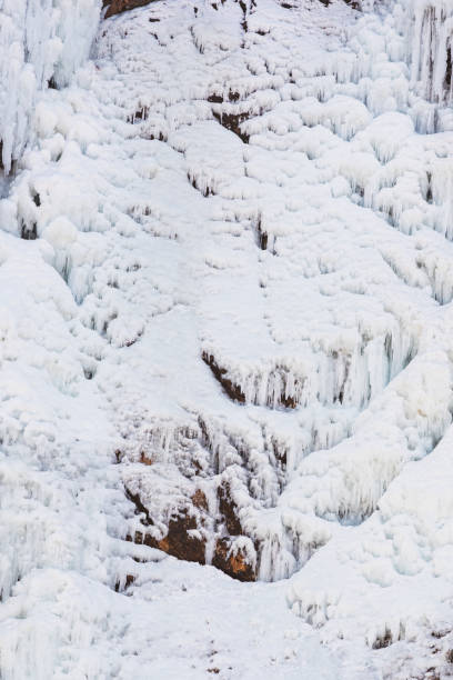 This photo shows the stunning scenery of winter. It features long icicles dangling from rough cliff edges and a stream, now turned to ice, winding through a valley blanketed in snow. The sunlight adds warmth to the chilly view, making the ice and snow glisten and showing off the plants peeking through the snow.