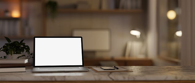 A white-screen laptop computer mockup on a white marble table in a cosy home office at night. home workspace concept. 3d render, 3d illustration