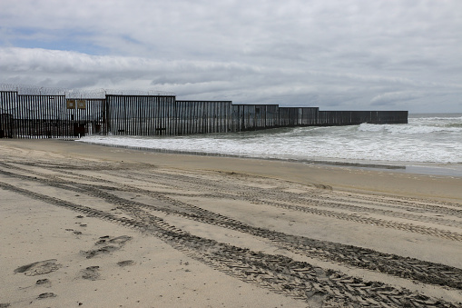 San Diego/Border Field Park, CA, USA - May 21, 2022. Photo taken from the beach, Playas de Tijuana, from the US side of the international border wall that separates San Diego, CA. from Tijuana, Mexico.  Footprints and border patrol vehicle tire tracks lead up to the steel wall.  The structure is topped with barbed wire and juts out into the Pacific Ocean where it ends.