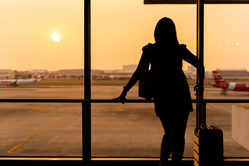 A woman is standing in front of a window, holding a suitcase. The light from the window is shining behind her, creating a backlit effect. The woman is the only person in the image.