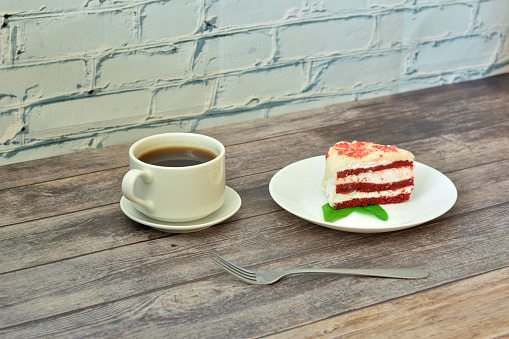 A plate with a piece of red velvet cake with mint leaves and a cup of bitter black coffee on a wooden table. Close-up.