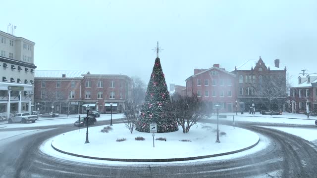 Small town America during white Christmas. Aerial reveal of snow covered roundabout in square with snow falling on Christmas tree during blizzard.