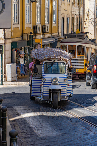 White, blue-striped three-wheeled tourist tuk tuk or Autorickshaw, going up a cobblestone street with tracks and tram rails in the Alfama neighborhood of Lisbon, followed by a yellow tram.