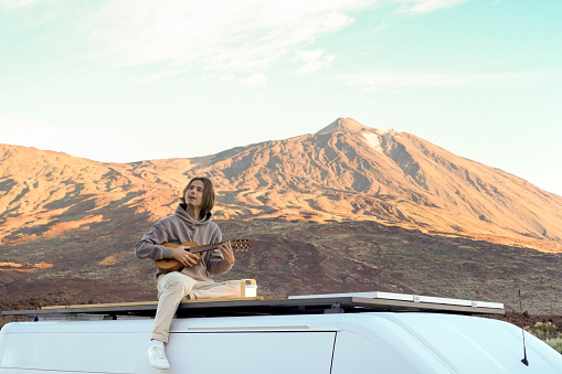 A young man sitting on top of a van in the picturesque Teide Mountain, playing a guitar.
