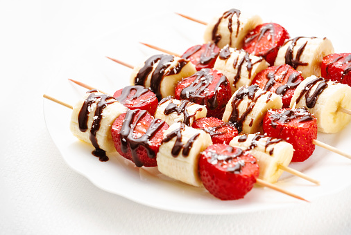Strawberries and bananas on skewers decorated with chocolate on a plate on a white background. Sweet snack. Healthy fresh dessert