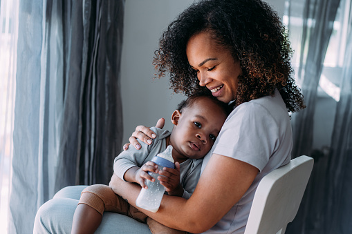 Portrait of a happy African-American woman holding and feeding her cute baby boy and enjoying motherhood at home. Shot of a cute little boy drinking milk from a baby milk bottle.
