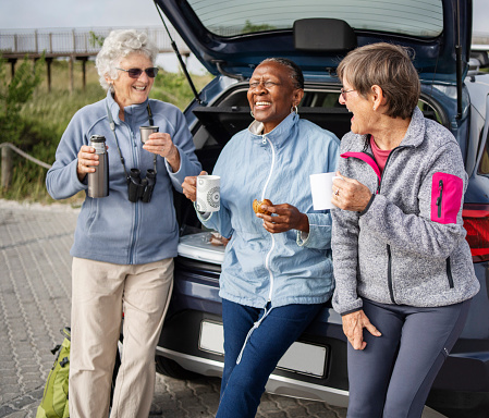 Laughing group of senior women eating snacks and drinking coffee while leaning against the trunk of their car during a road trip together