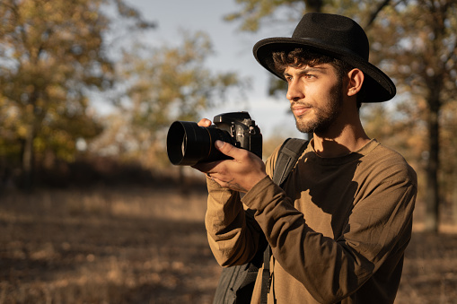 Portrait of travel photographer wearing hat with digital camera making photo of the nature at sunset. Copy space