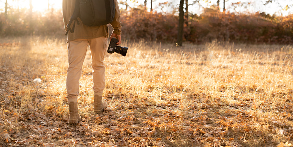 Unrecognizable man with backpack holding camera in autumn forest, nature travel and hiking concept. Banner. Copy space