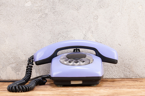 Vintage blue rotary dial telephone on wooden table in front gray concrete wall background.