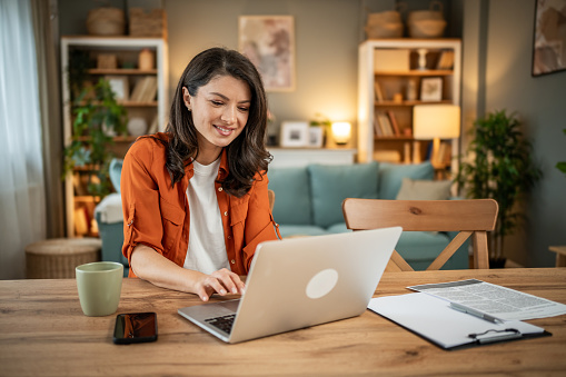 A satisfied young woman, a freelancer, is sitting at the dining room table and working on a laptop