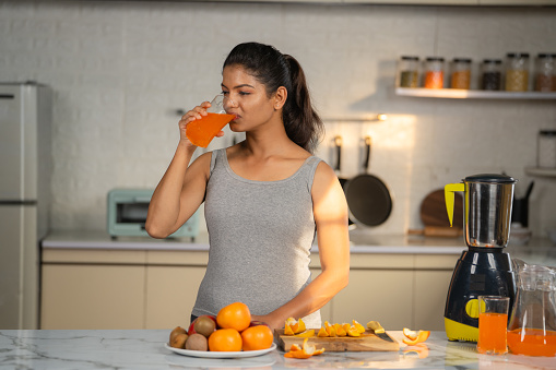 Young Indian woman in active wear drinking orange juice by drinking at kitchen - concept of healthy or wellness and morning fitness routine