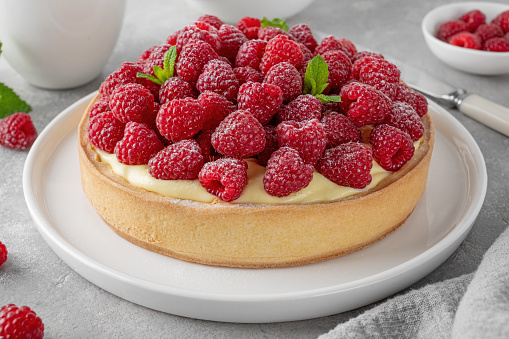 Delicious raspberry tart with vanilla custard cream sprinkled with powdered sugar on a white plate on a gray concrete background. Summer dessert