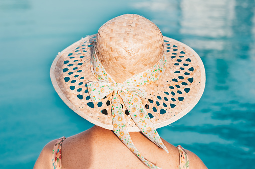 Rear view of caucasian senior woman in swimsuit and straw hat enjoying freedom and vacation sitting at the outdoor swimming pool.