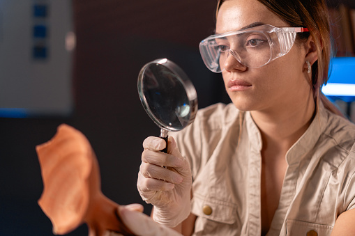 Close-up of a female archaeologist scientist examining an ancient artifact through a magnifying glass while working late at night in the office. Copy space
