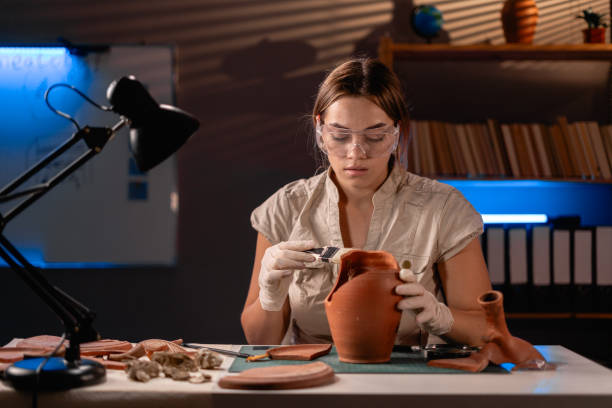 オフィスで深夜に作業する女性考古学者、研究用のブラシで古い壺を掃除する - tools for construction ストックフォトと画像