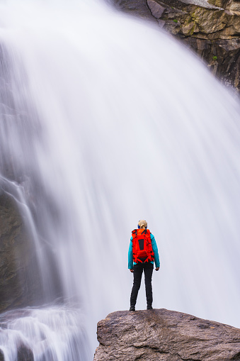 rear view of hiking woman with backpack standing on rock close to waterfall in the mountains