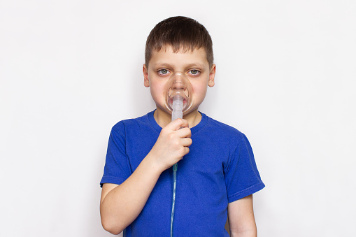 Portrait of a little boy who uses a nebulizer for inhalation and inhales medicine through a mask, isolated on a light gray background. Cough or bronchial asthma treatment concept