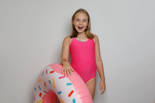 Joyful child girl with pink rubber inflatable ring swimming pool float standing on white studio wall background