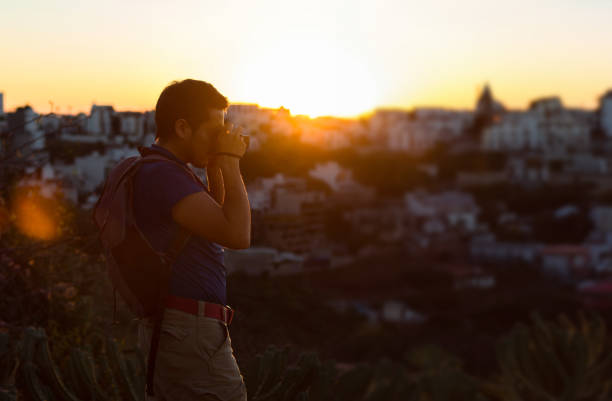 A man in a plaid shirt sits on rocky terrain, using binoculars to scan a green valley below. The scene captures outdoor adventure, nature exploration, and the quiet anticipation of discovery at dusk.