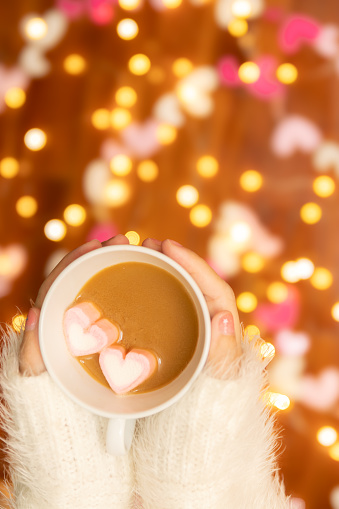 woman hand with sweater holding coffee cup with heart shape marshmallow on top, defocus bokeh light decorate for Valentine's Day celebration at background with copy space. romantic love relationship