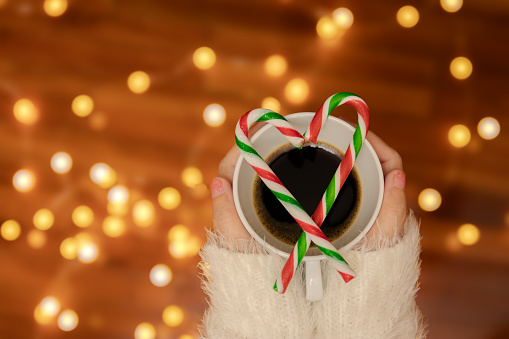 woman hand with sweater holding coffee cup with candy canes in the shape of heart on top, defocus bokeh light decorate for Valentine's Day celebration at background with copy space. romantic love