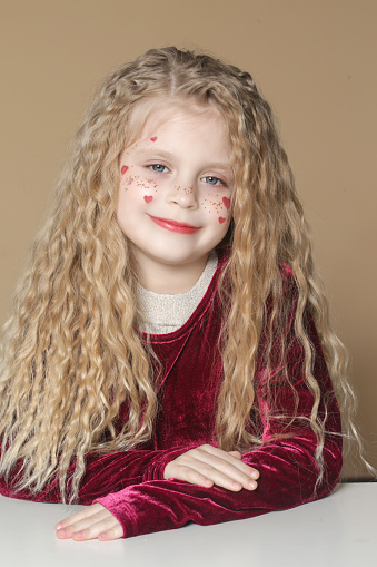 Portrait of cute little girl in red dress with long blonde hair and glitters and red hearts on the face