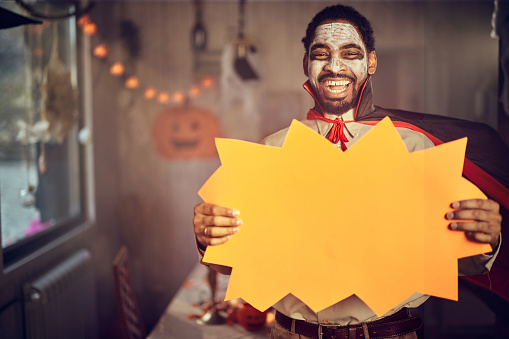 Happy African American man in count Dracula costume holding empty placard at home and looking at camera. Copy space.