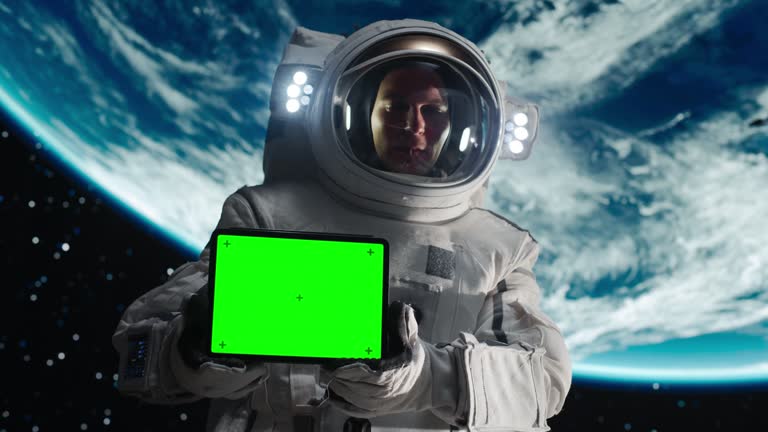 Portrait of a Young Male Astronaut Posing in a Space Suit During a Spacewalk on a Satellite or a Space Station. Spaceman Holding Tablet Computer with Green Screen Display in Horizontal Orientation