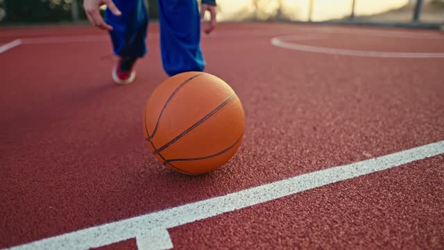 Shooting close up a blonde girl in a sports uniform picks up an orange basketball from the red floor that is bouncing on a street court in the morning