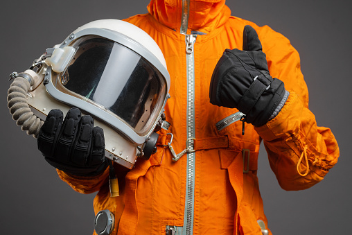 Astronaut is standing in the space suit and helmet in hand and showing a thumbs up gesture sign on the gray background. Space tourist.