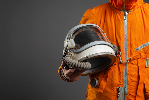 Astronaut is standing in the space suit and helmet in hands on the gray background with copy space.