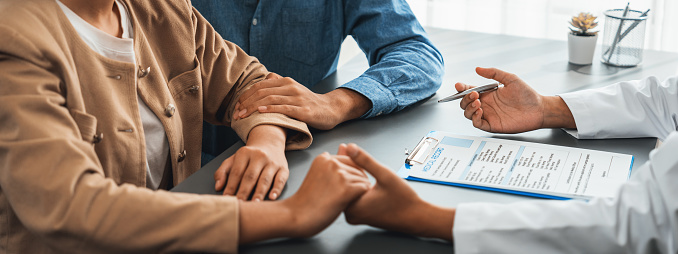 Doctor providing compassionate healthcare consultation while young couple patient holding hand, comfort each other after infertile report. Reproductive and medical fertility consulting. Neoteric