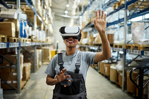 Happy African American female worker having fun while using virtual reality simulator in distribution warehouse.