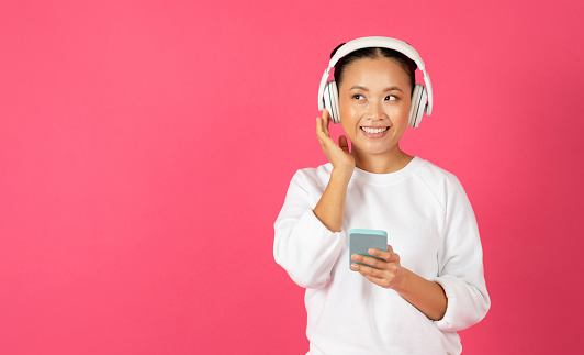 Music App. Cheerful Asian Woman Listening Her Favorite Songs With Smartphone And Wireless Headphones, Happy Smiling Young Korean Lady Standing Against Pink Studio Background, Copy Space