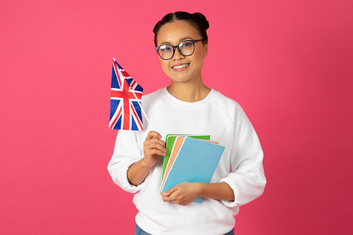 Beautiful young asian student woman holding flag of United Kingdom and books, happy korean female recommending international studies and exchange programs, standing against bright pink background