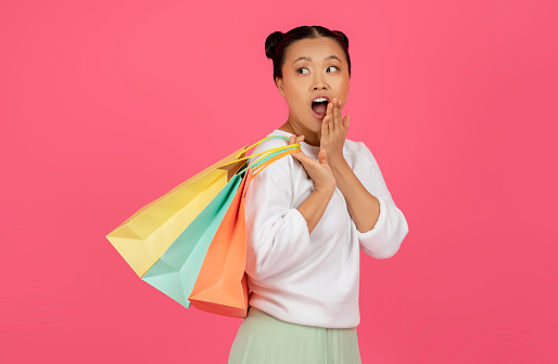 Excited asian female with shopping bags touching her face in surprise, shocked korean lady cannot believe huge sale, posing isolated on pink studio background, pretty woman buying on discount